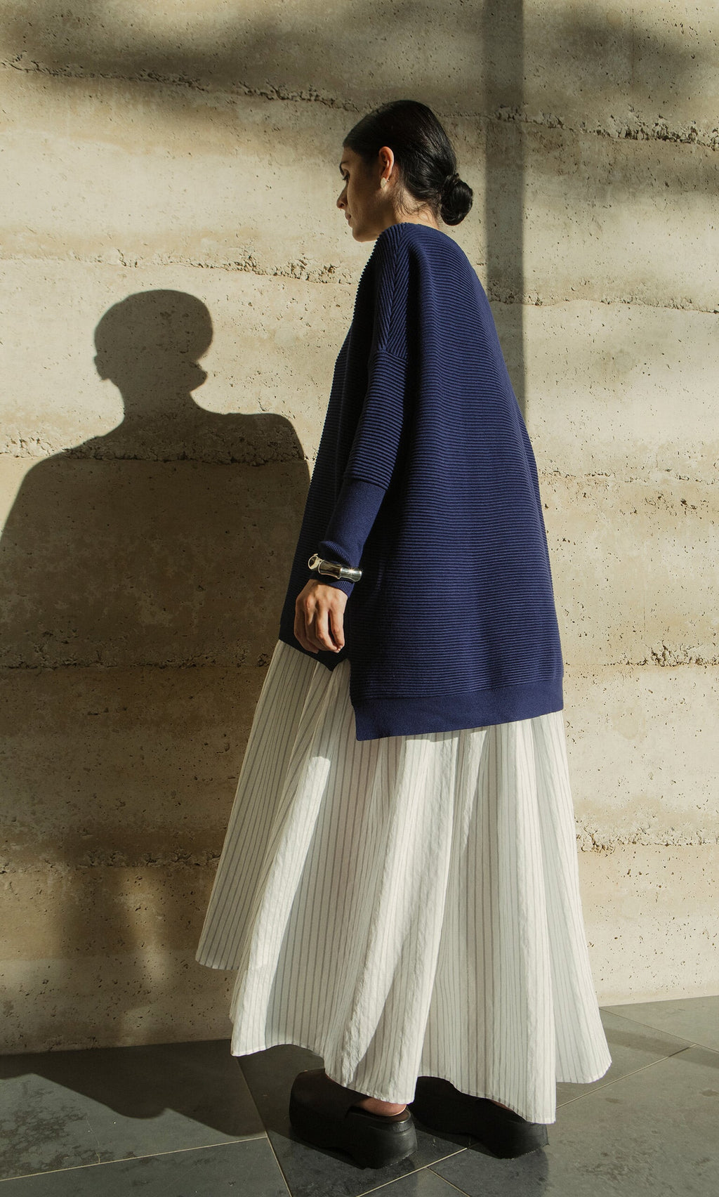 Plümo- Woman in the Jack Sweater - Navy stands sideways in sunlight, wearing a long white skirt against a beige wall.