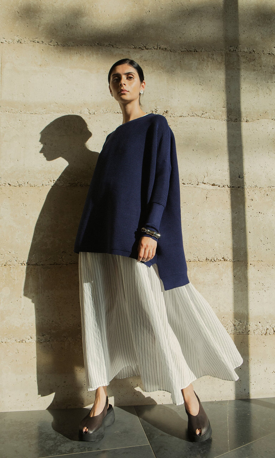 Plümo- Woman in Jack Sweater - Navy with dropped shoulders and white pleated skirt stands in sunlight, casting a shadow.