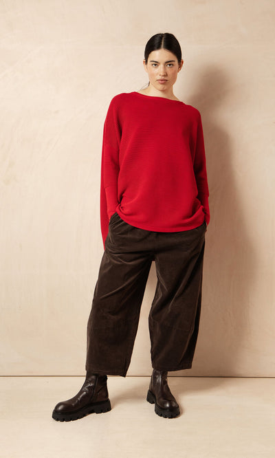 Plümo Woman in a red sweater and Anise Pants with elasticated waist, hands in pockets, on beige background.