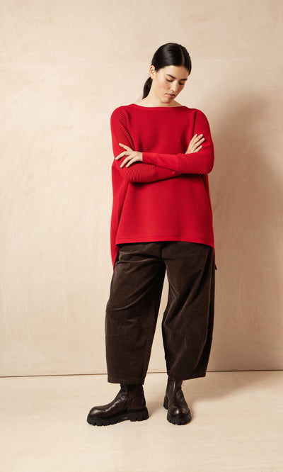 Plümo- Woman in a red sweater, Anise Pants, and black boots stands with arms crossed, looking down.