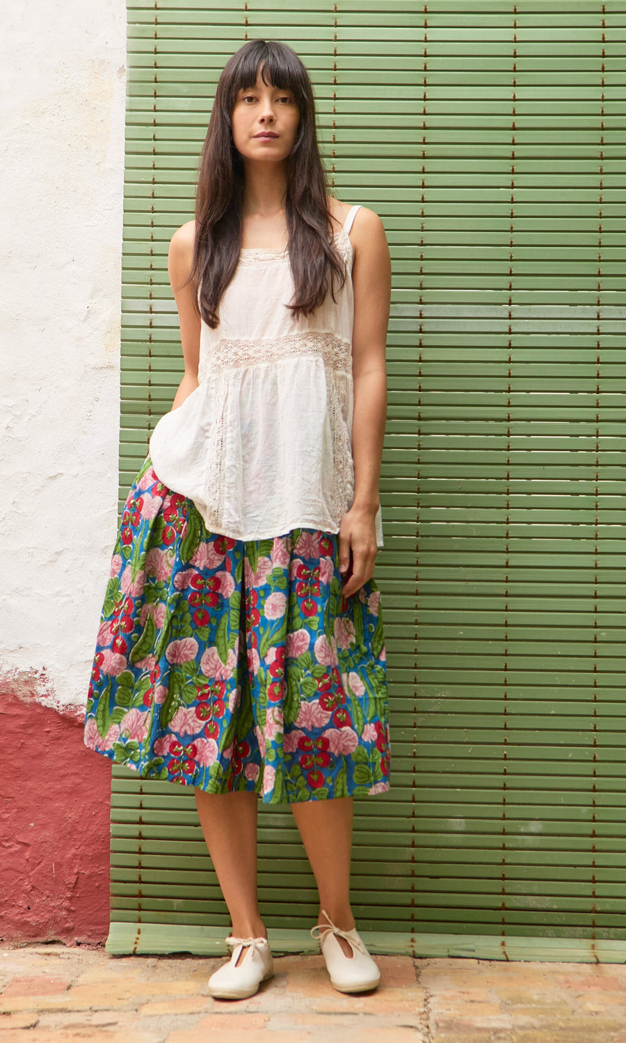 Plümo- Woman in a white sleeveless top and Jardin Skirt stands before a green bamboo blind.