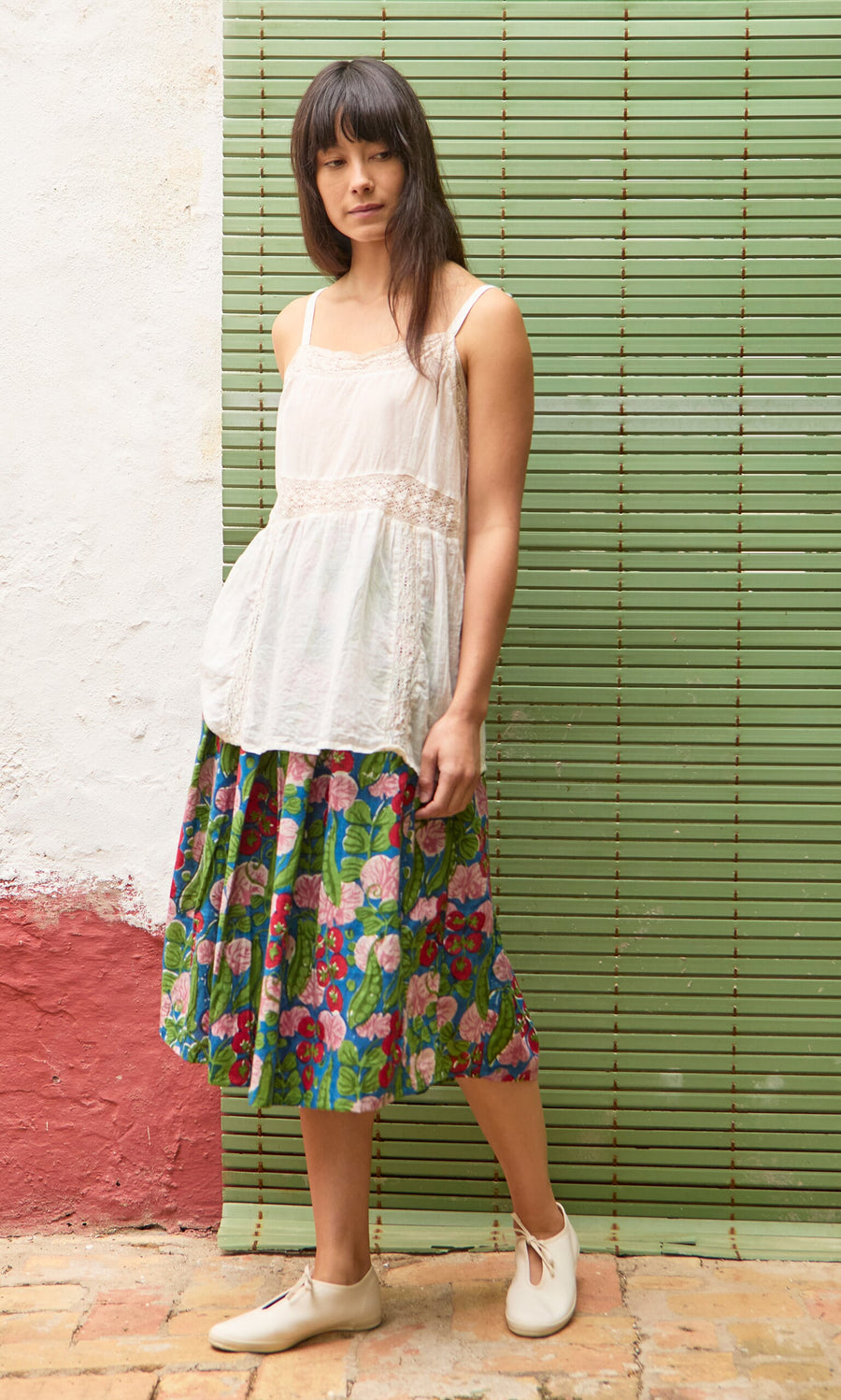 Plümo- Woman in a white sleeveless top and the Jardin Skirt stands before a green bamboo curtain.