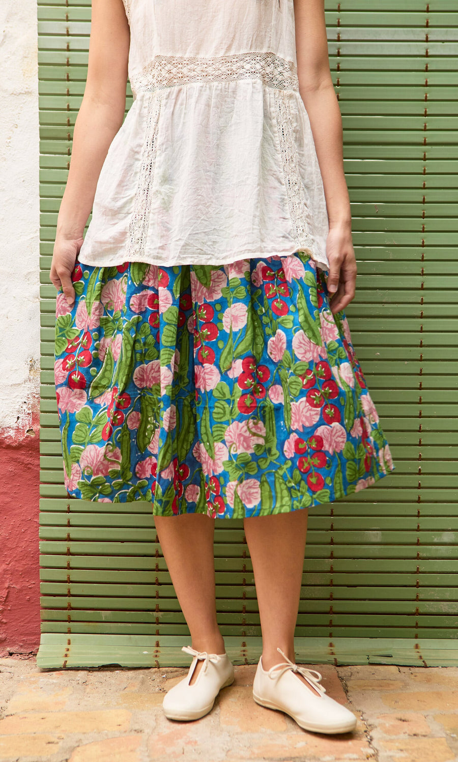 Plümo- A person in a white blouse and the Jardin Skirt stands before a green bamboo screen.