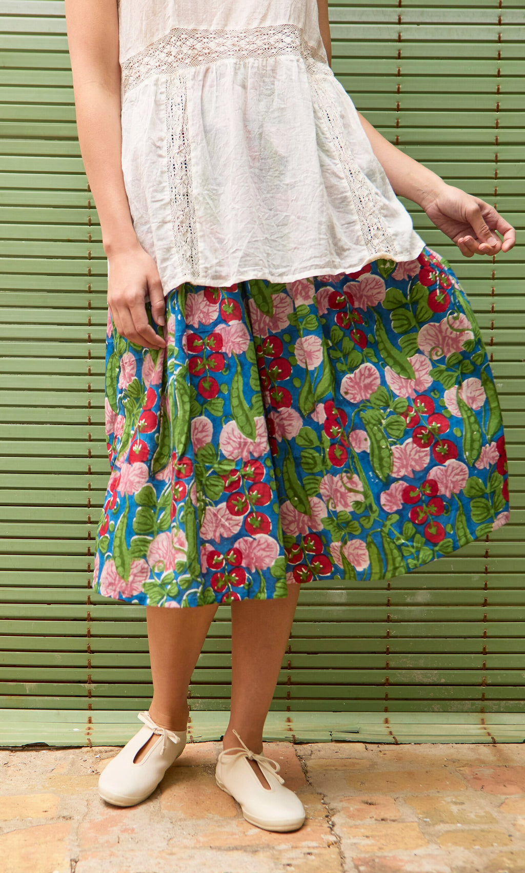 Plümo- A person wears the Jardin Skirt with a shirred waist and a white blouse, standing before a green striped background.