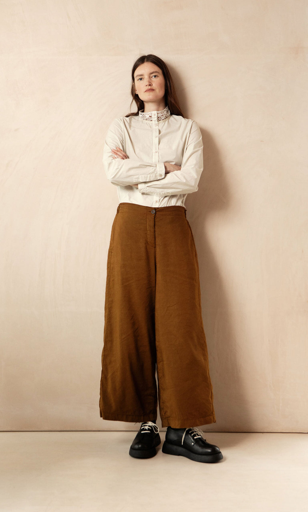 Plümo- A woman in a white blouse and Kapur Pants with a flattering silhouette stands with arms crossed by a beige wall.
