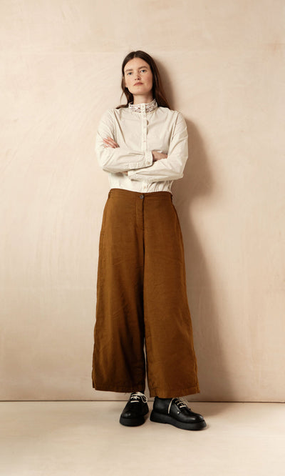 Plümo- A woman in a white blouse and Kapur Pants with a flattering silhouette stands with arms crossed by a beige wall.