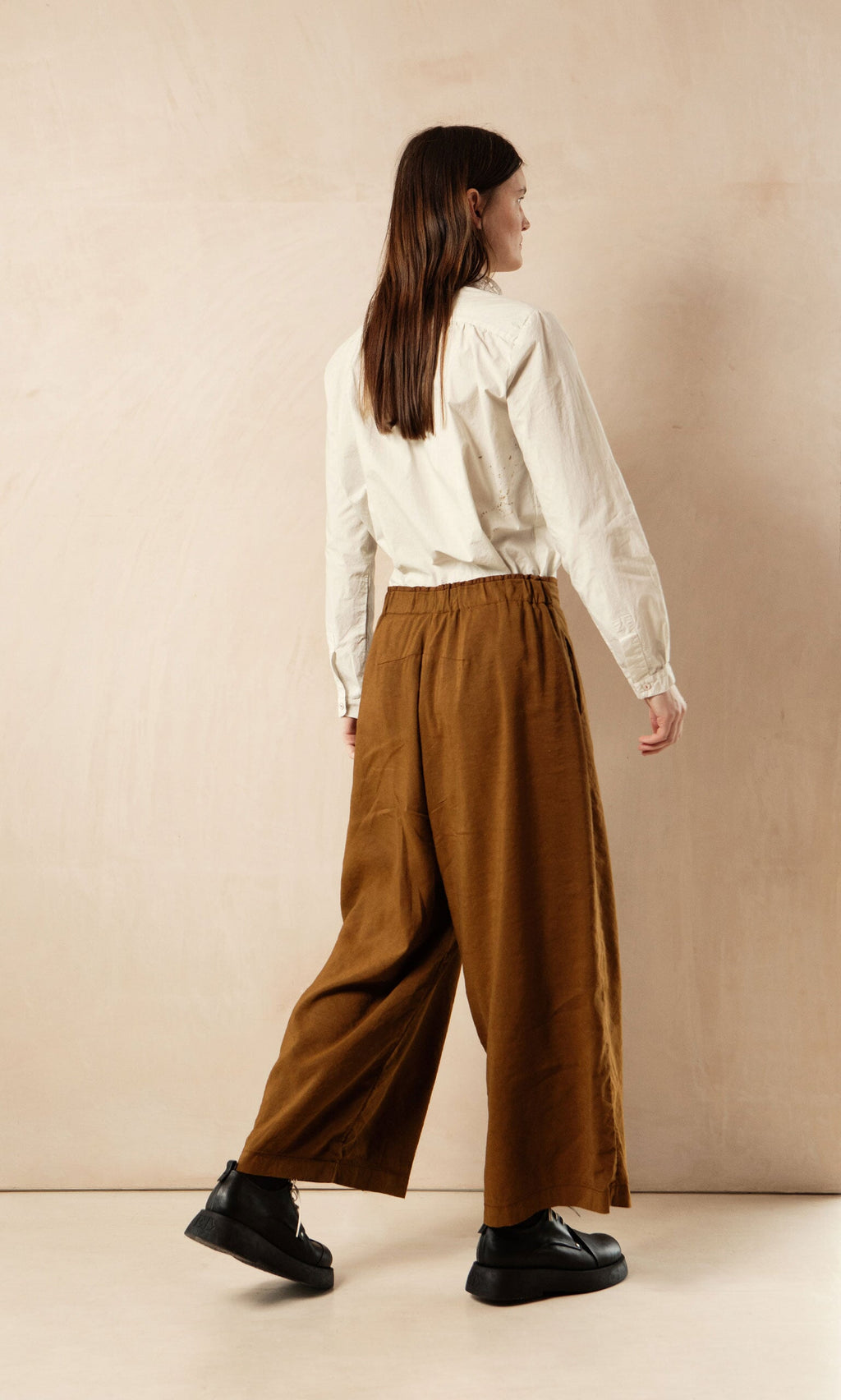 Plümo- Someone in a cream blouse and brown Kapur Pants with a flattering silhouette stands against a beige background.