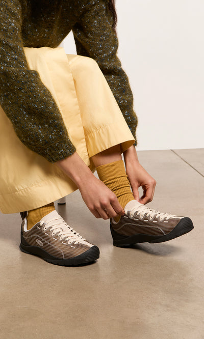 Plümo  Person tying beige Walking Shoes, wearing yellow socks and skirt, sitting on a bench.