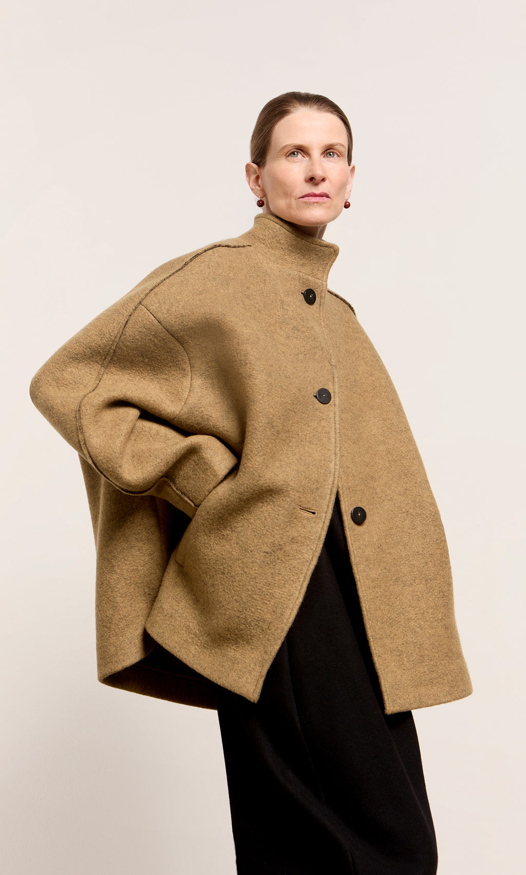Plümo  Someone wears the Kilmore Coat, an oversized Italian wool design with black buttons, against a plain backdrop.