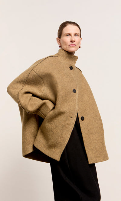 Plümo  Someone wears the Kilmore Coat, an oversized Italian wool design with black buttons, against a plain backdrop.