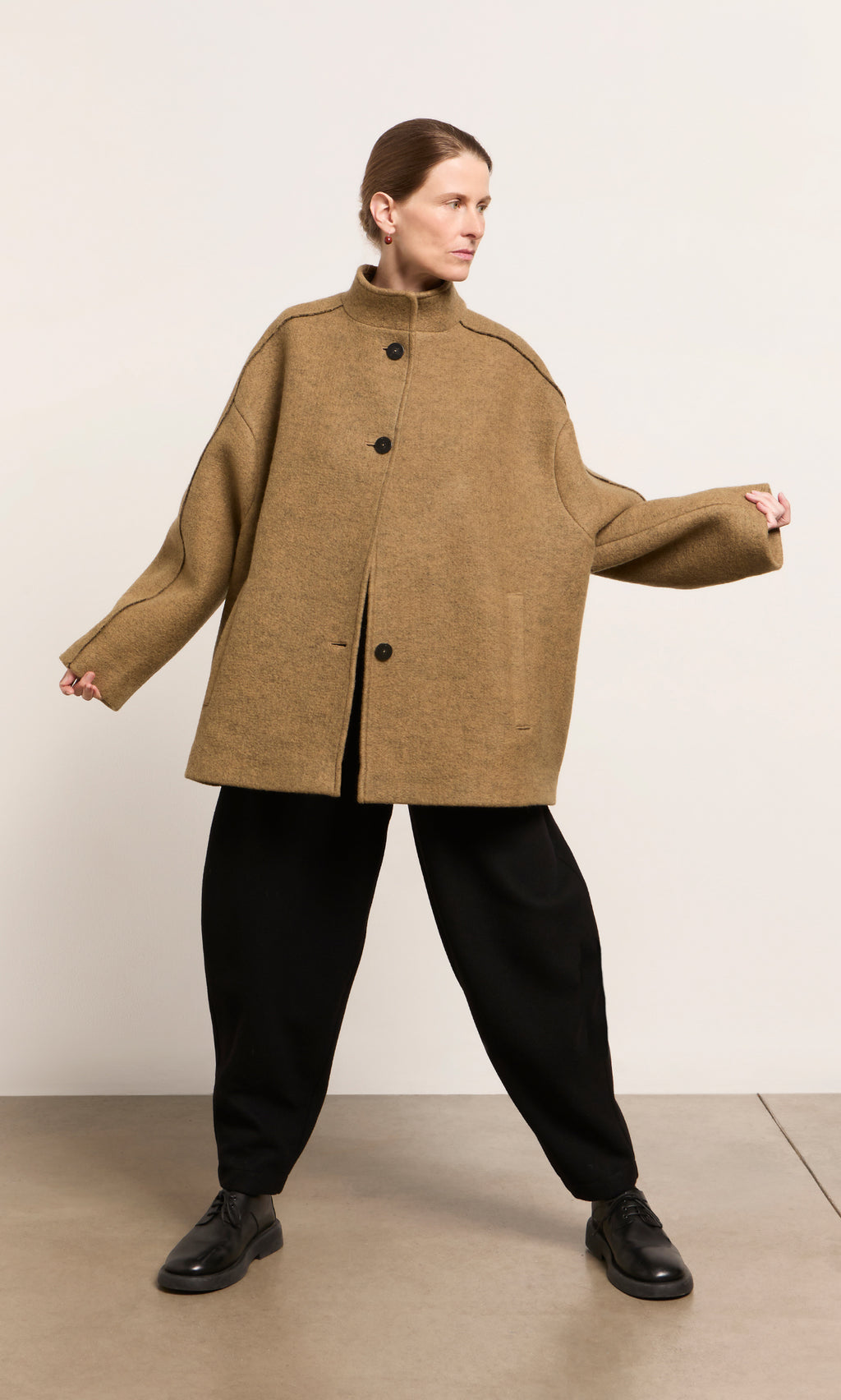 Plümo  Person in a Kilmore Coat acorn brown, black pants, and shoes stands with arms out against a plain background.