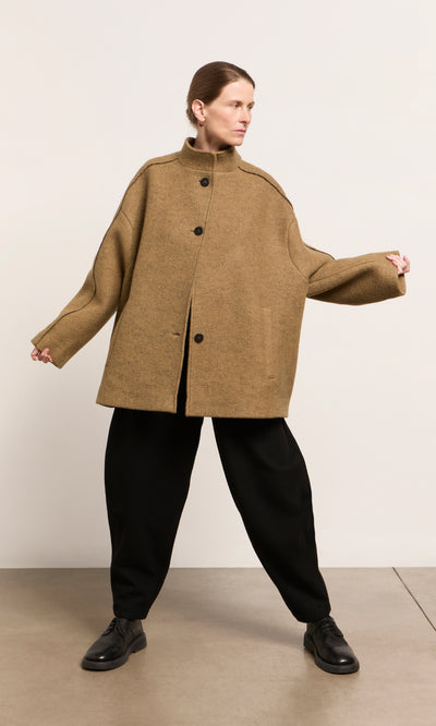 Plümo  Person in a Kilmore Coat acorn brown, black pants, and shoes stands with arms out against a plain background.