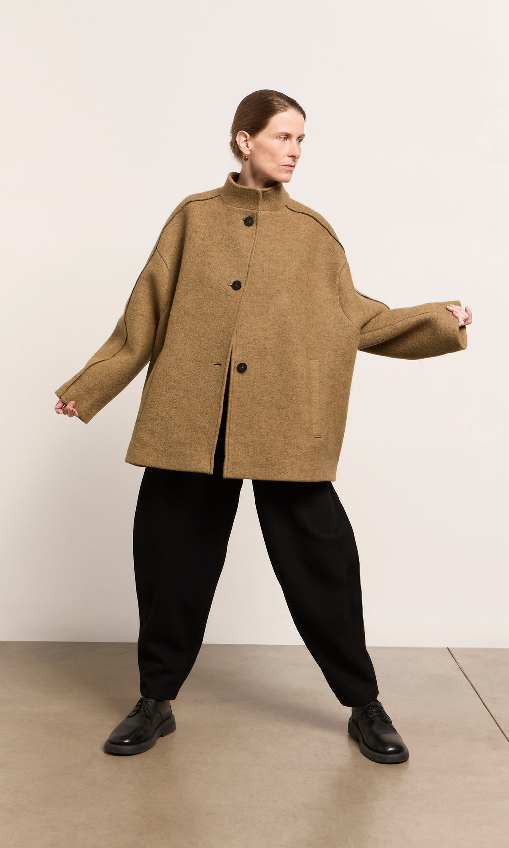 Plümo  Woman wears the oversized Kilmore Coat in acorn brown with black pants, arms outstretched against a plain background.