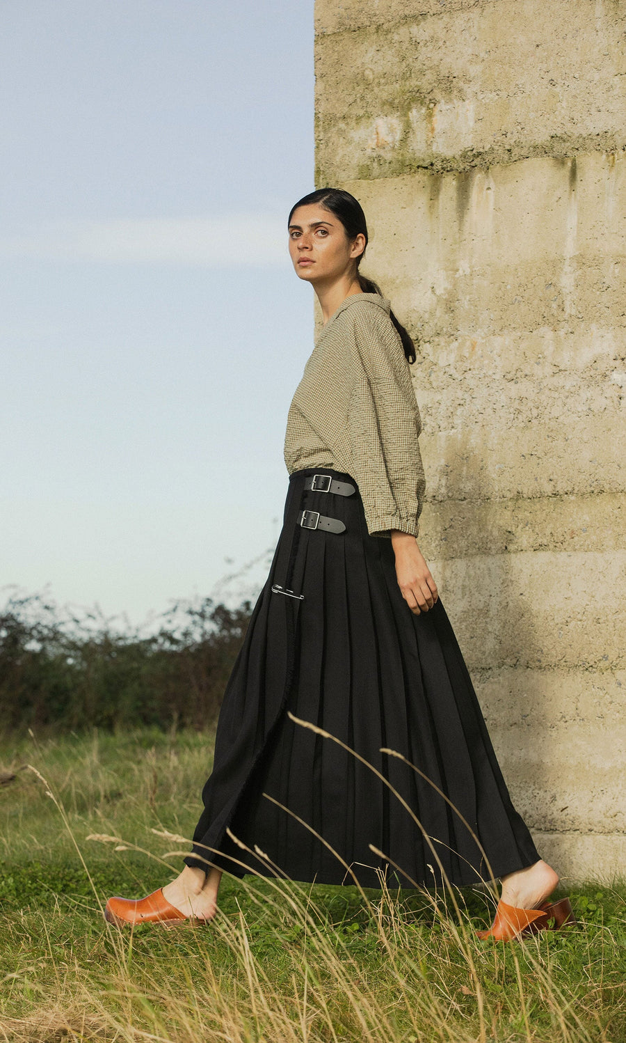 Plümo- Woman wears a green blouse and Storm Kilt as she walks outdoors by a concrete wall with grass and sky behind.
