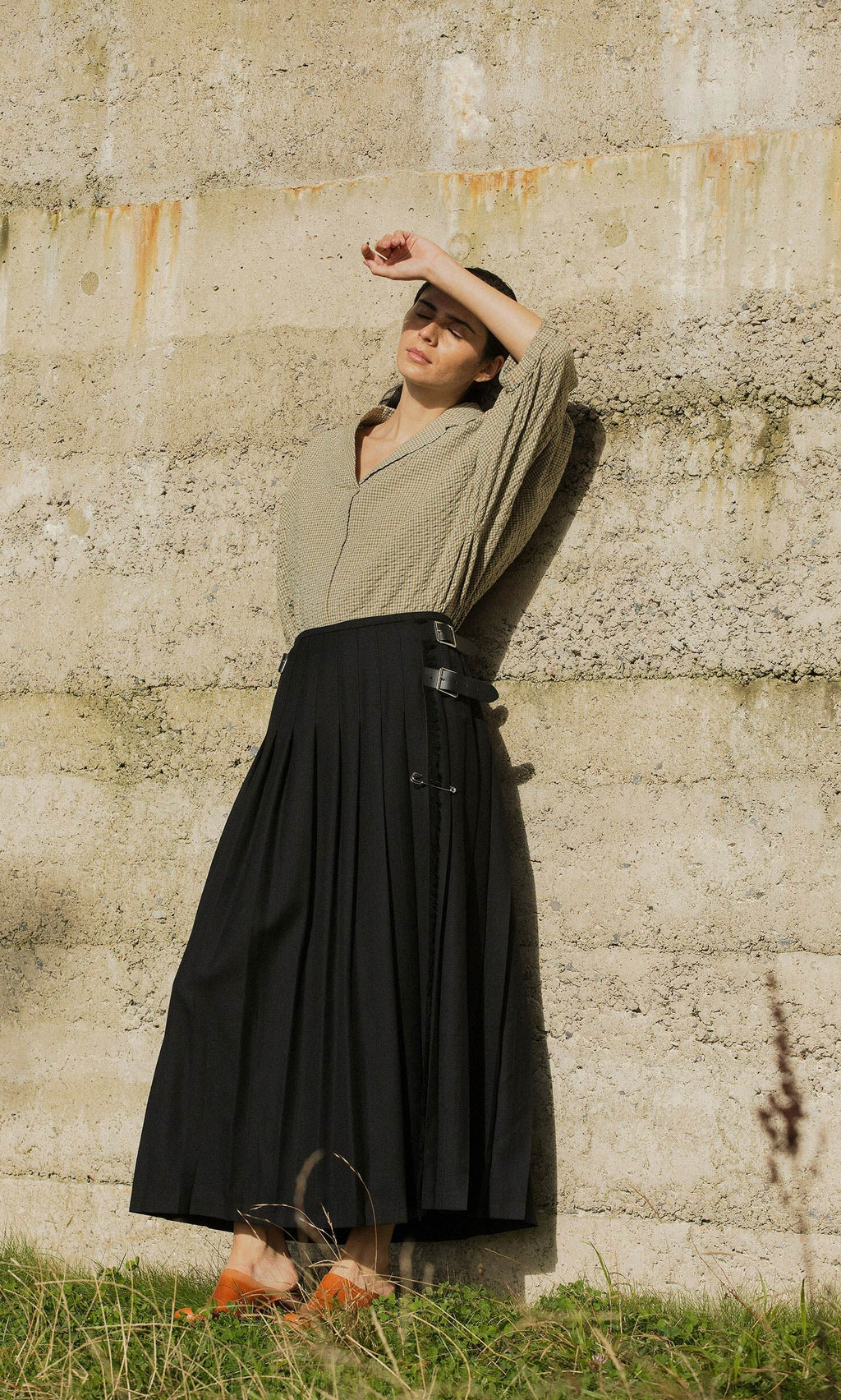 Plümo- Woman in beige top and Storm Kilt stands by a textured wall, shading her face from the sun with her hand.