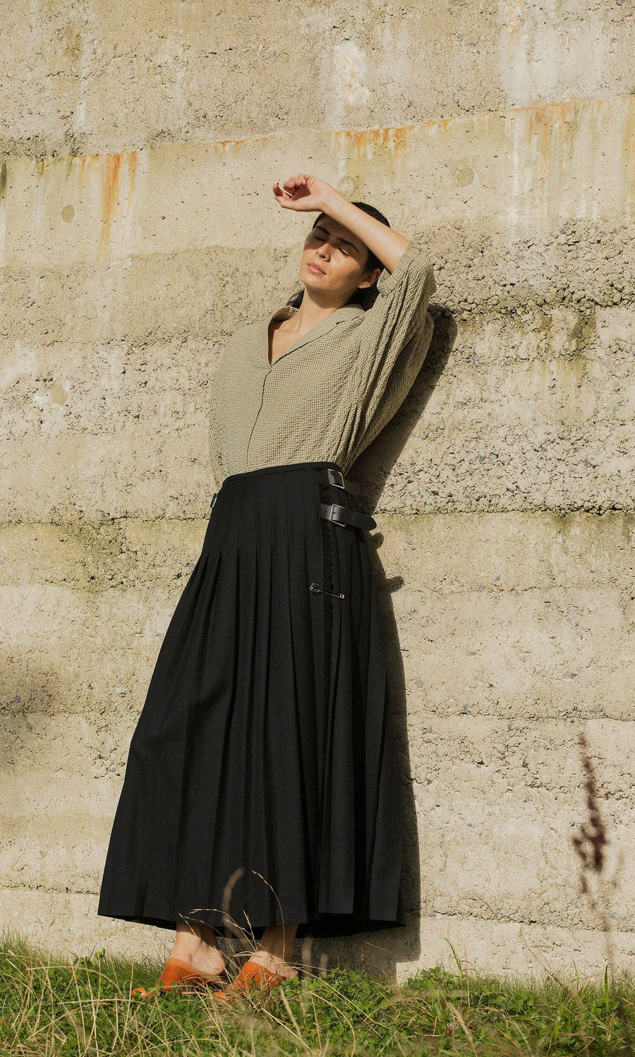 Plümo- Woman in beige top and Storm Kilt stands by a textured wall, shading her face from the sun with her hand.