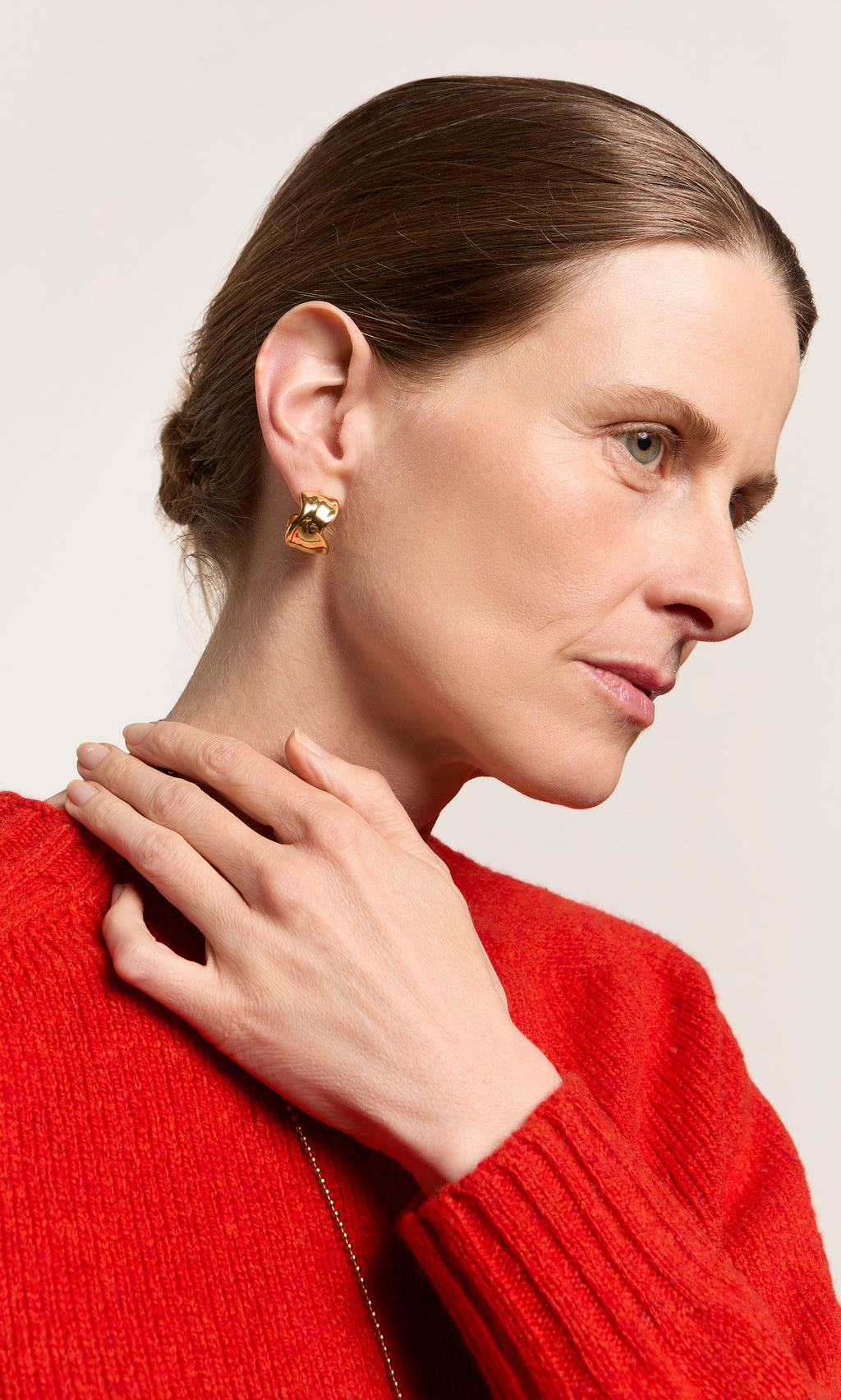 Plümo  Woman in red sweater touches neck, showcasing Melted Hoops earrings with hair back on a plain background.