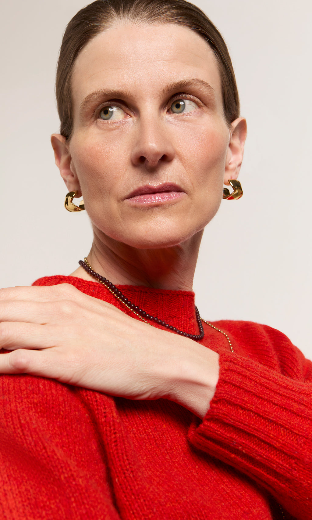 Plümo  Woman in a red sweater wearing Melted Hoops earrings and a necklace, looking to the side with her hand on her shoulder.