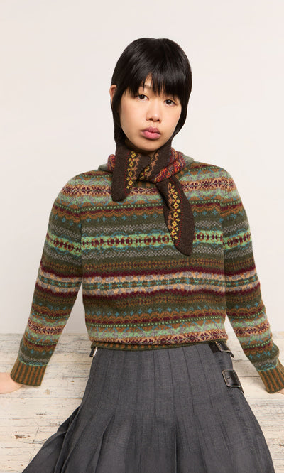 Plümo  Individual wearing the Lenie Necktie, a patterned sweater, and pleated skirt sits before a light background, gazing ahead.