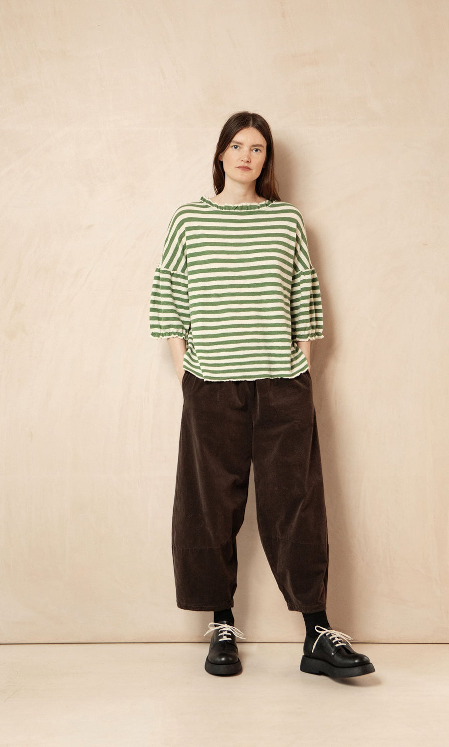 Plümo- Woman wears the Lina Frill Blouse, a green striped relaxed fit top, and loose brown pants, standing by a beige wall.