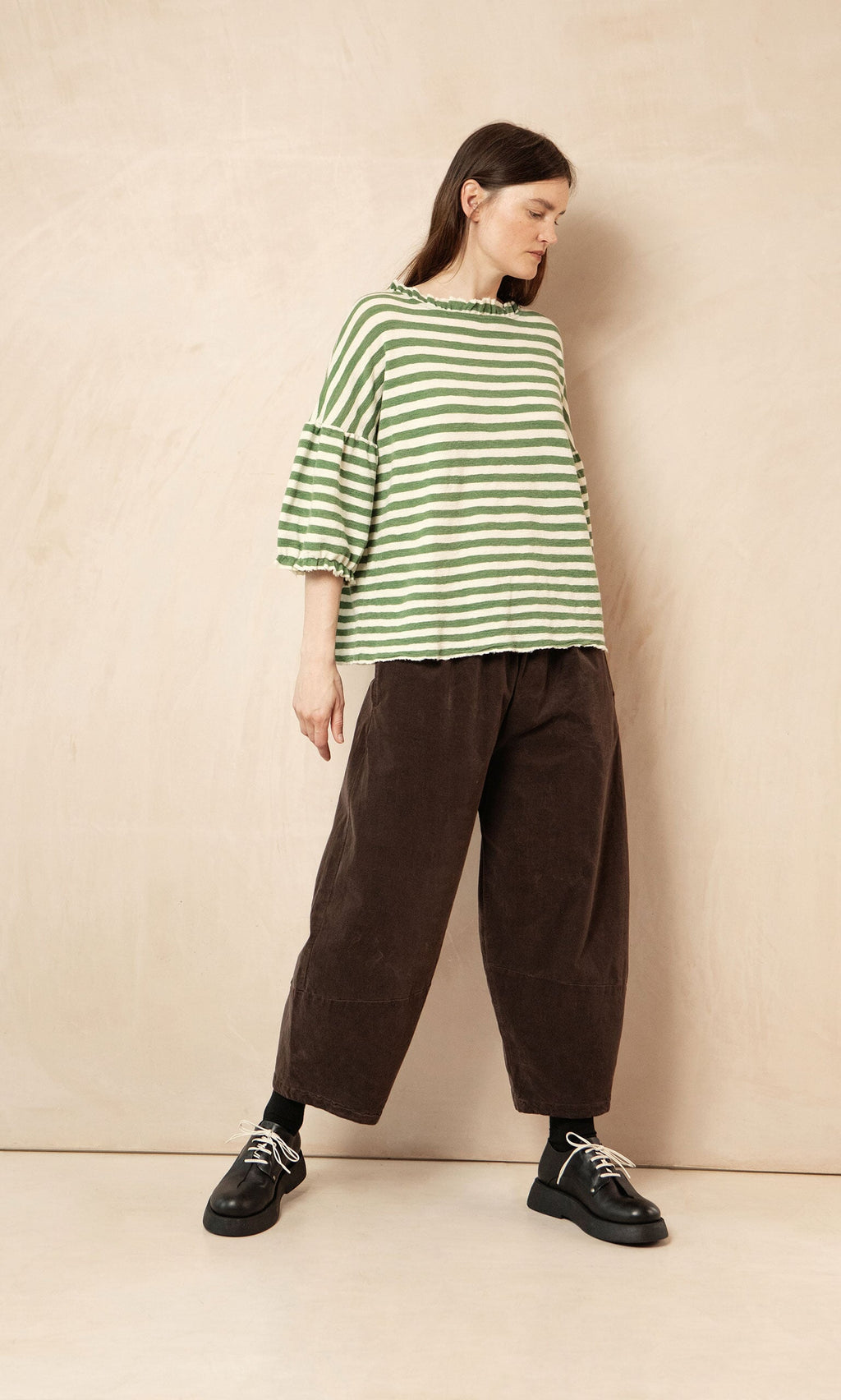 Plümo- Woman in a Lina Frill Blouse and loose brown pants stands against a beige wall, looking to her left.