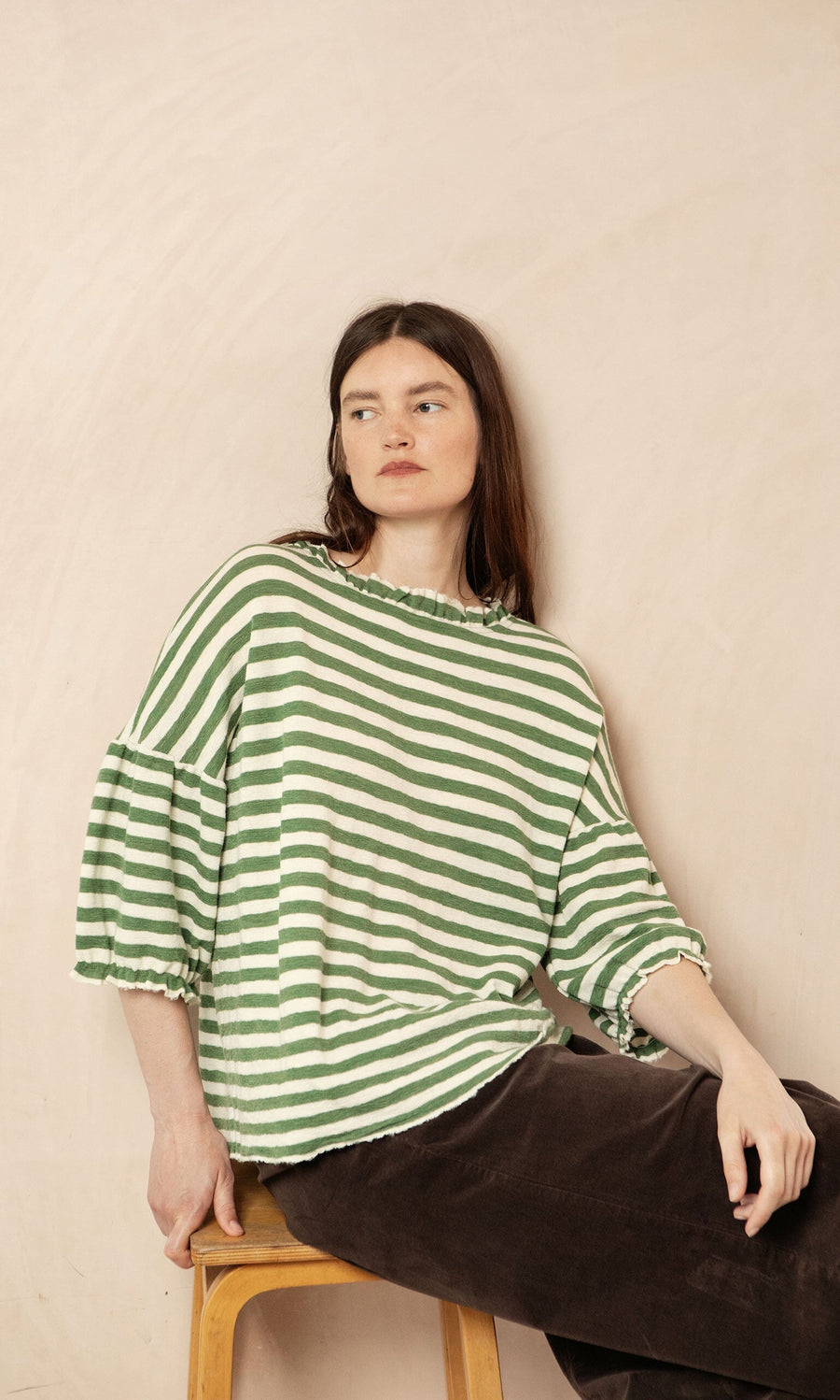 Plümo- Woman with long brown hair wears the Lina Frill Blouse in green and white stripes, seated on a stool.