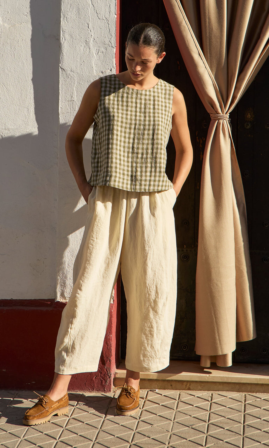 Plümo- Wearing the Linda Top - Olive Gingham and cream wide-leg pants, person stands by a doorway with tan curtains.