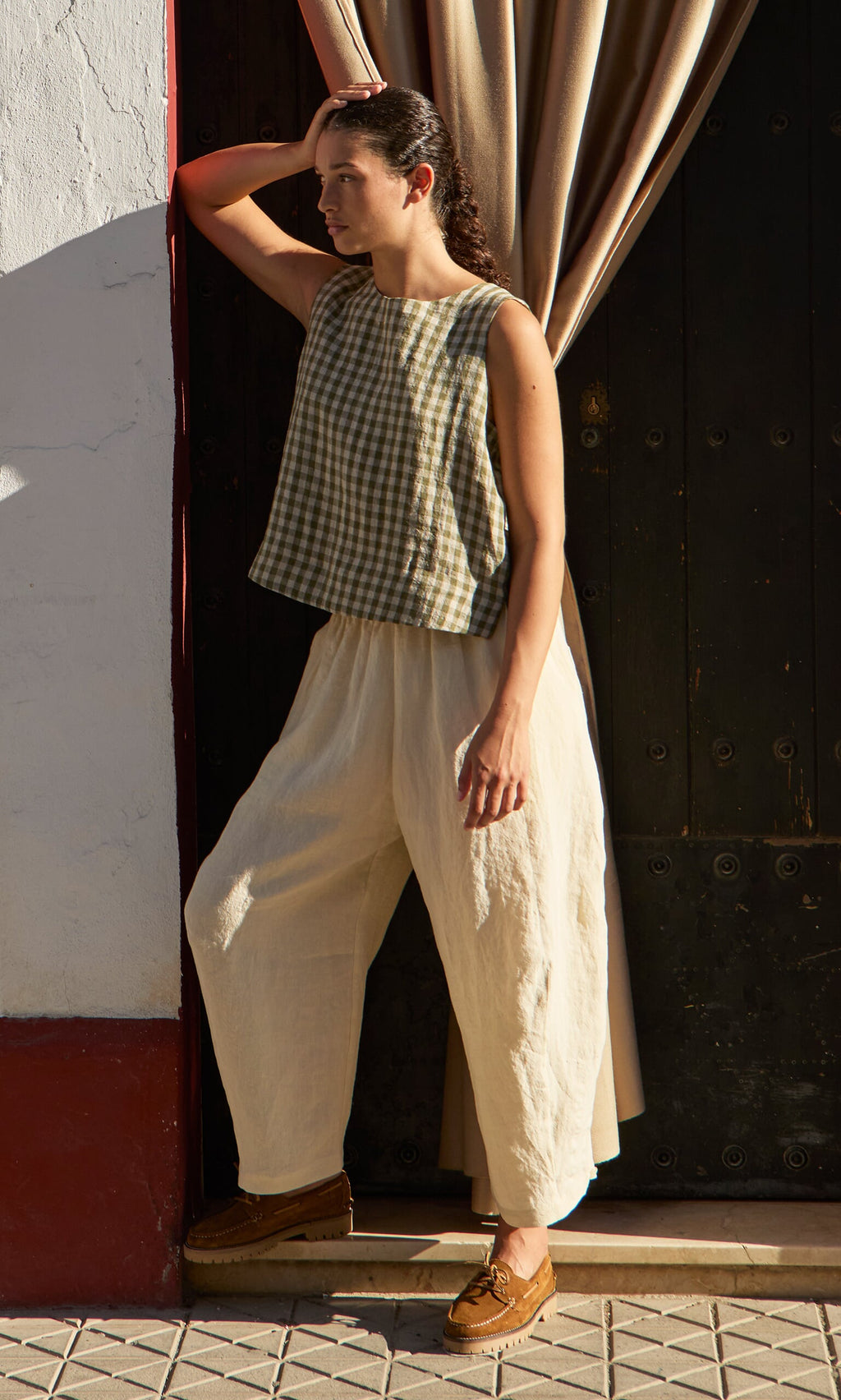 Plümo- Person in the Linda Top - Olive Gingham and loose cream pants stands by a doorway, one arm raised to their head.