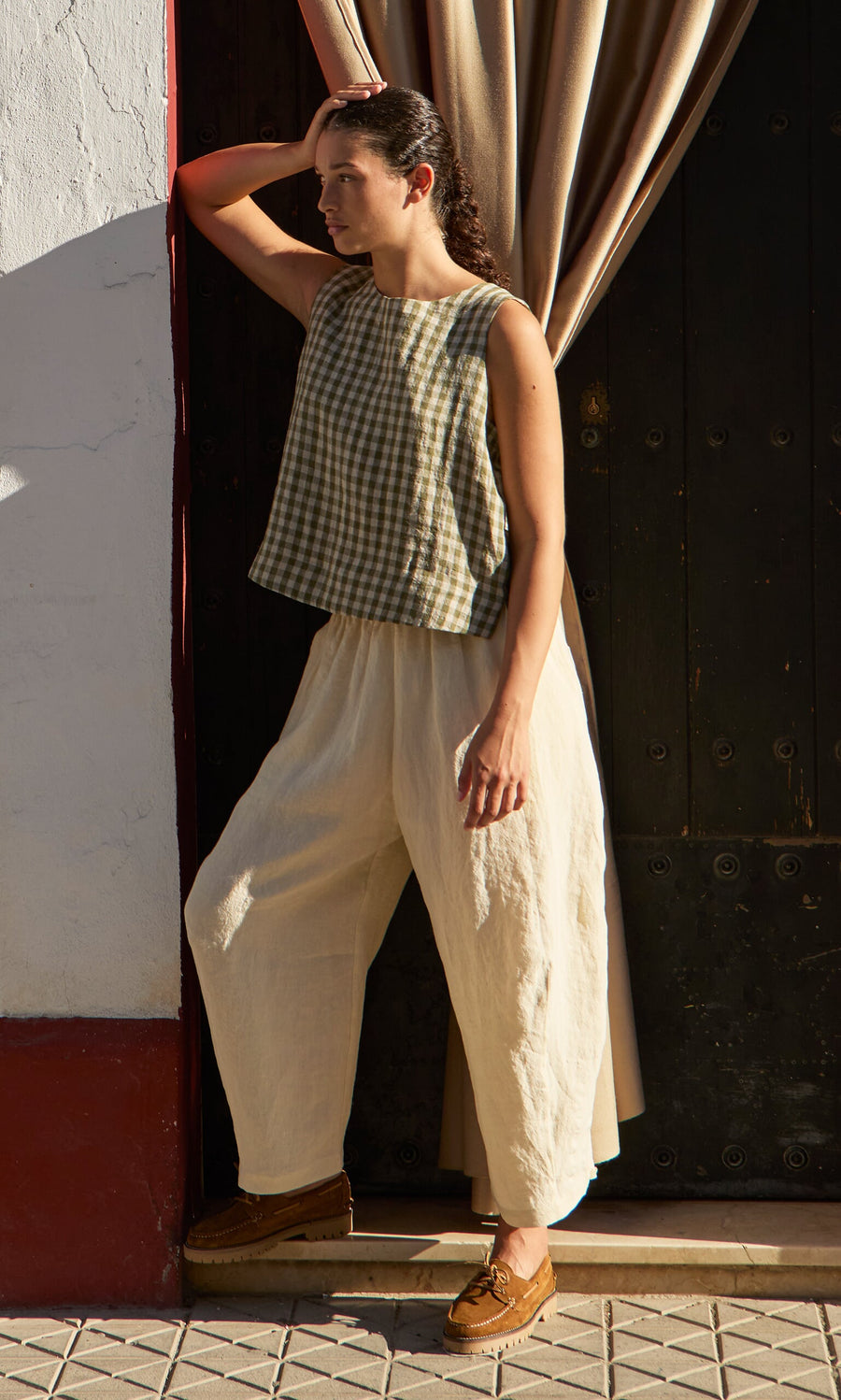 Plümo- Person in the Linda Top - Olive Gingham and loose cream pants stands by a doorway, one arm raised to their head.