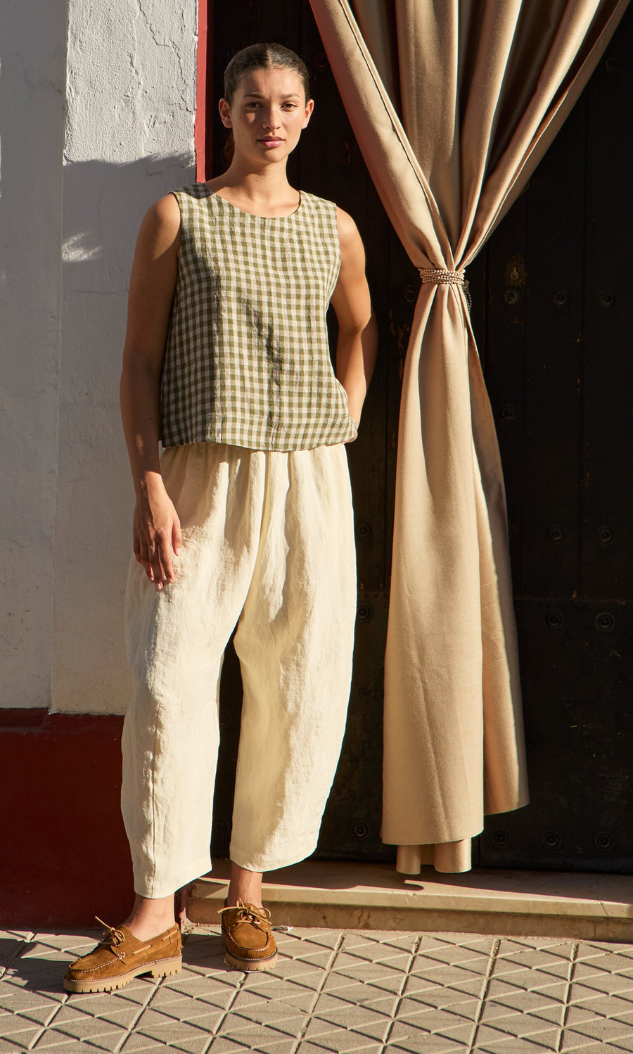 Plümo- Woman in the Linda Top - Olive Gingham, loose cream pants, and brown shoes stands outdoors by a curtain and door.