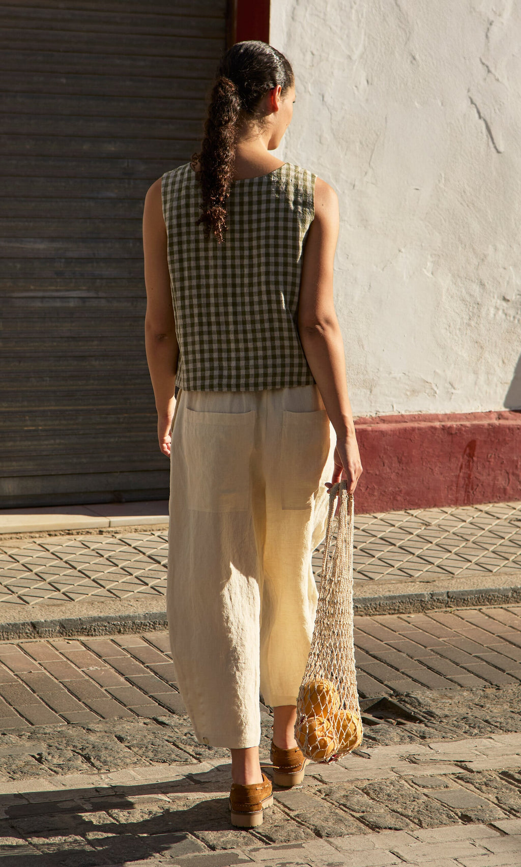 Plümo- A woman in the Linda Top - Olive Gingham and cream pants carries a net bag of oranges on a sunny street.