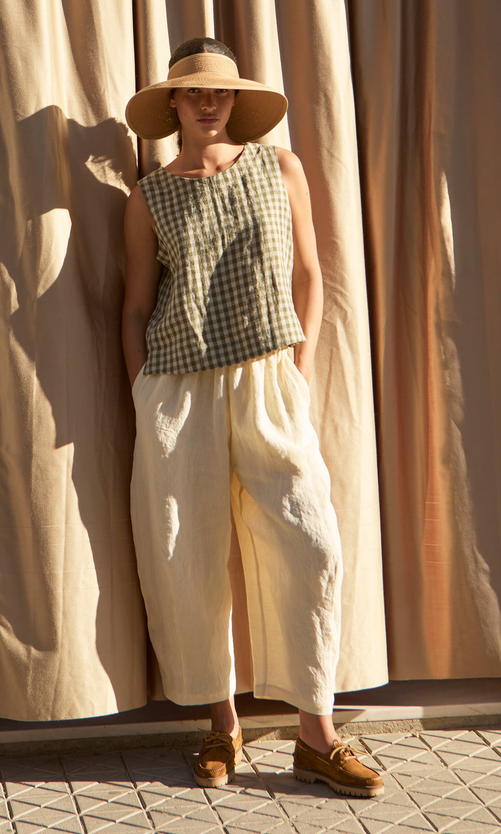 Plümo- Person in a wide-brim hat, Linda Top - Olive Gingham, loose cream pants, and brown shoes stands by beige curtains.