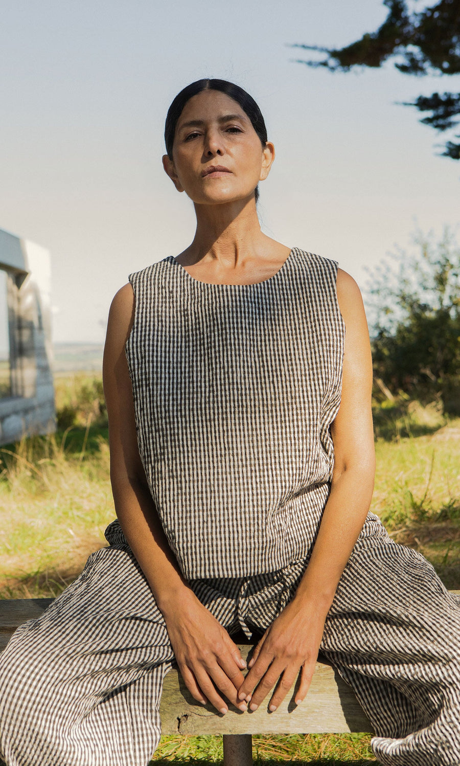 Plümo- A woman in the Linda Top - Gingham with a scoop neck sits outdoors, confidently gazing at the camera.