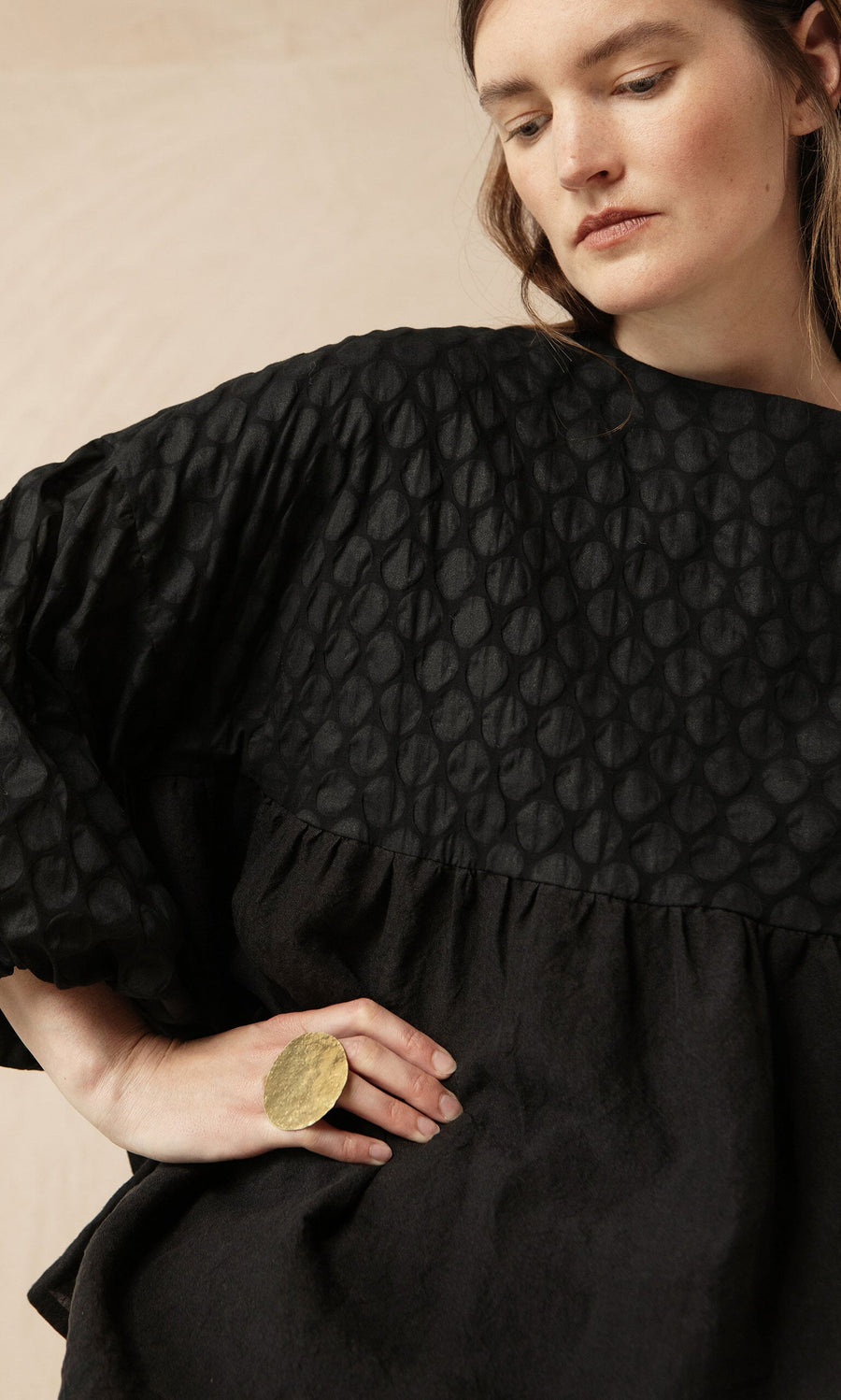 Plümo- Woman in the Tuppence Blouse and textured black dress poses with a large gold ring, hand on hip, looking downward.