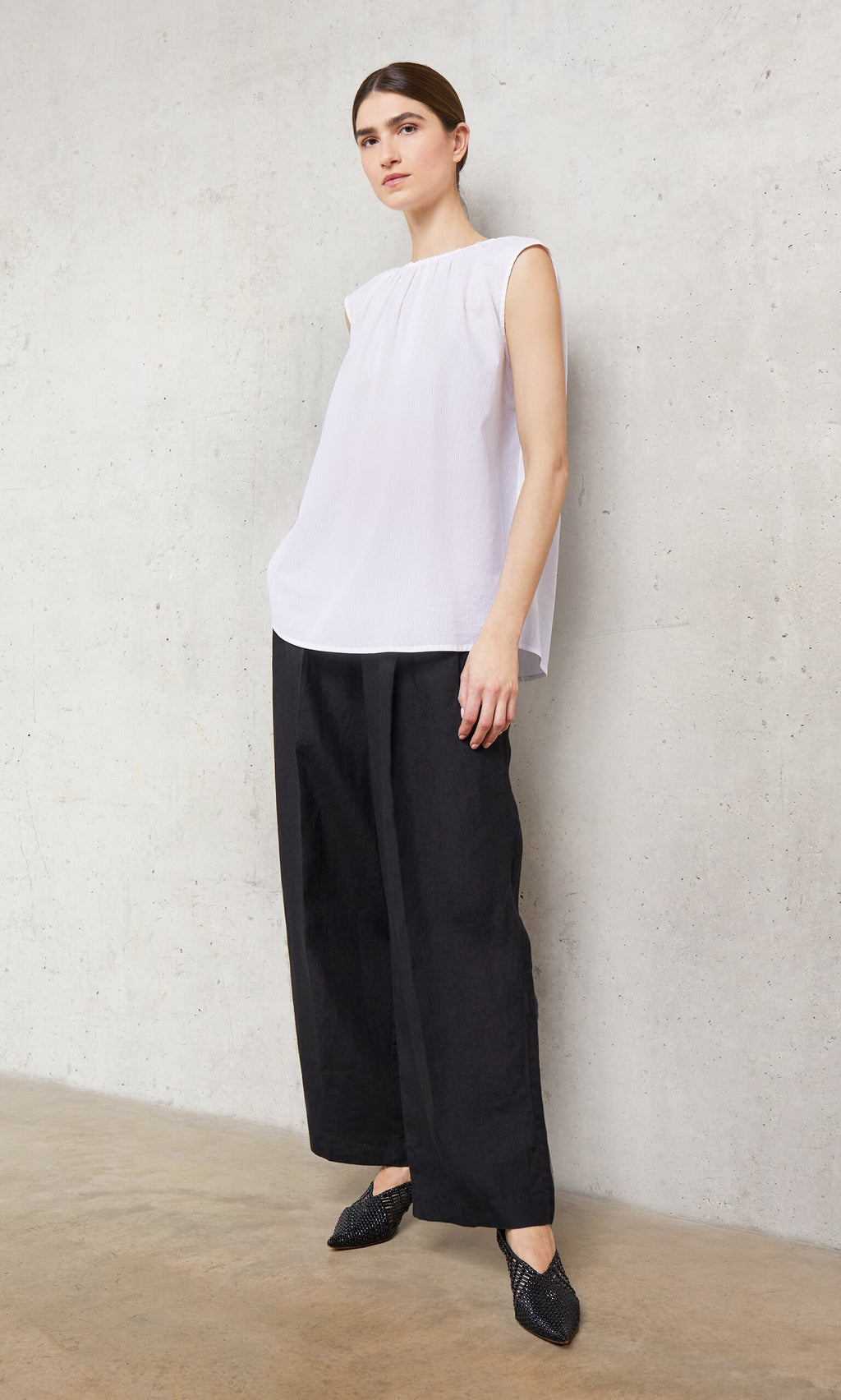 Plümo- Woman in a white Marie Top with side slits, wide-leg black pants, and black shoes stands by a light concrete wall.