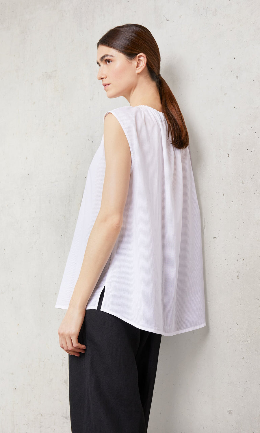 Plümo- Woman with brown hair in a low ponytail wearing the Marie Top - White and black pants, standing sideways.