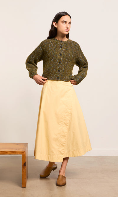 Plümo- A person in a green sweater, Marigold Wrap Skirt, and brown clogs stands indoors by a wooden bench.