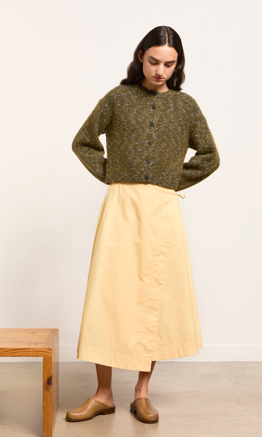 Plümo- Woman wearing a green cardigan, Marigold Wrap Skirt, and tan clogs, standing indoors by a wooden bench.