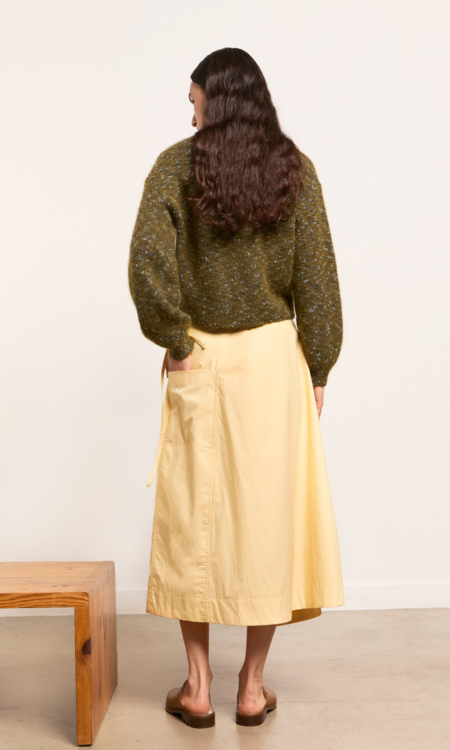Plümo- A person with long brown hair in a green sweater and Marigold Wrap Skirt stands by a wooden bench, facing away.