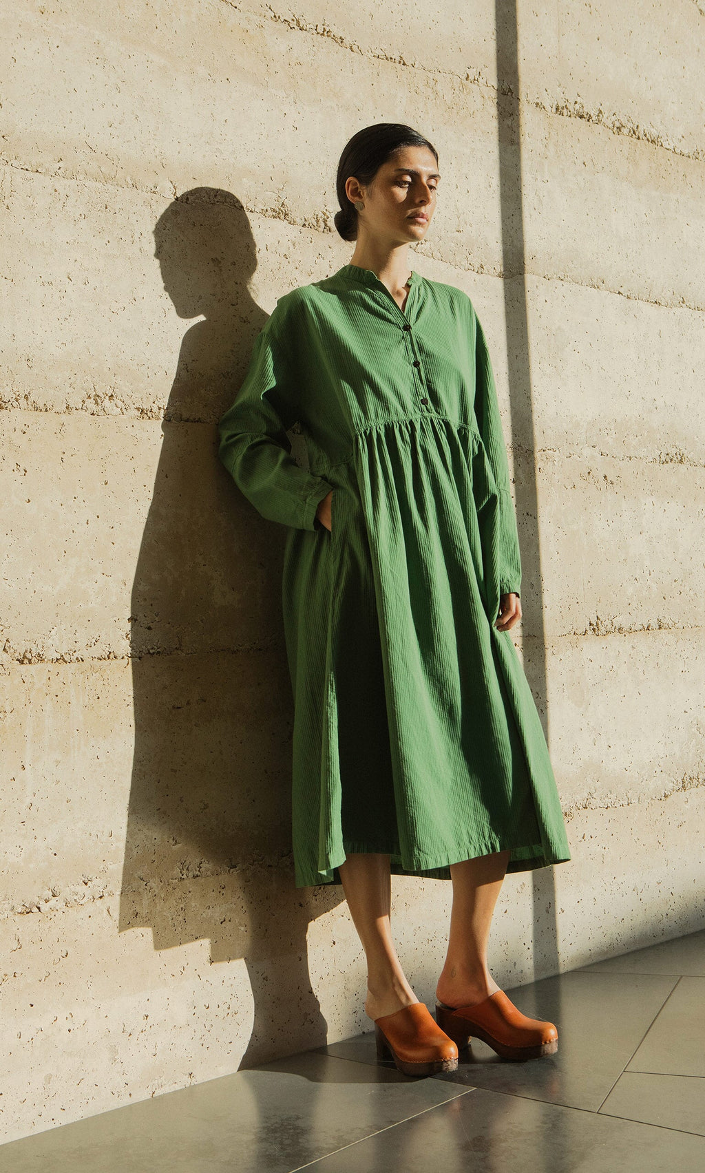 Plümo- Woman in the Marisol Dress, featuring bracelet-length sleeves, stands by a sunlit wall casting a shadow.