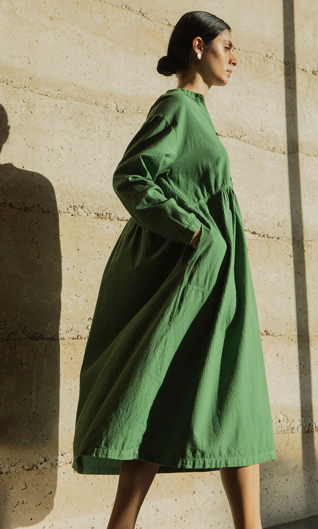 Plümo- A woman in the Marisol Dress, an emerald cotton style with bracelet-length sleeves, stands by a sunlit beige wall.