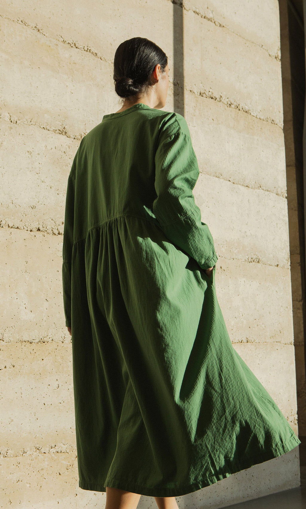 Plümo- A person in a Marisol Dress with bracelet-length sleeves stands outdoors facing a sunlit beige concrete wall.