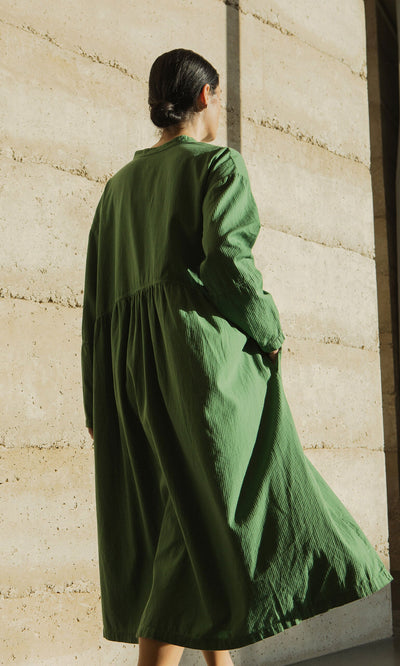 Plümo- A person in a Marisol Dress with bracelet-length sleeves stands outdoors facing a sunlit beige concrete wall.