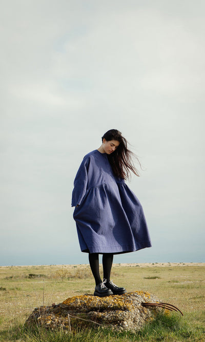 Plümo- Woman wearing the Matilda Dress with a full skirt stands on a rock in a grassy field, wind blowing her hair.