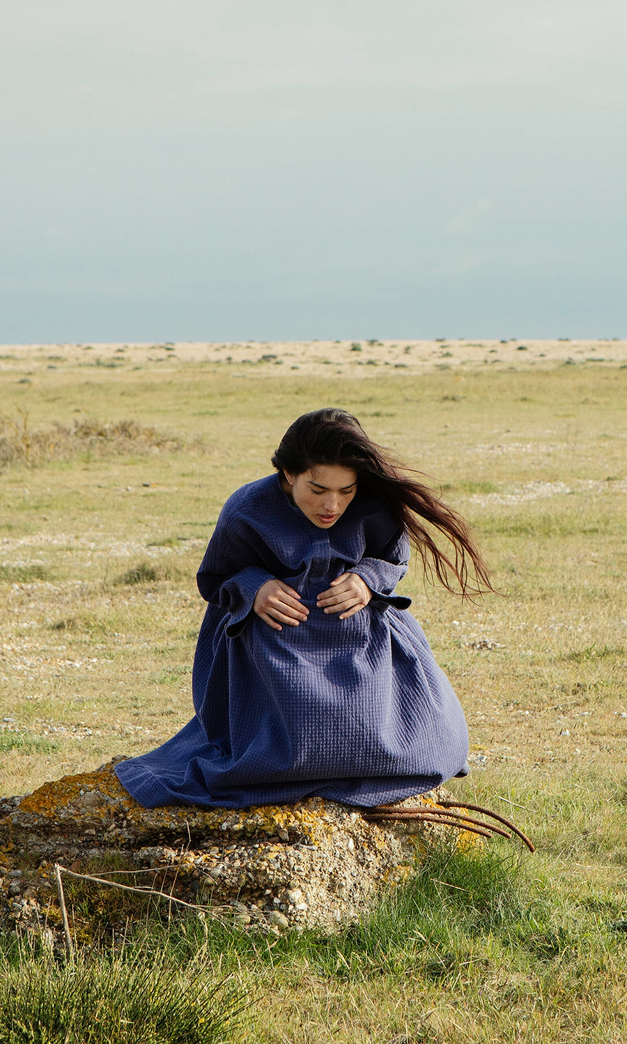 Plümo- A woman in the Matilda Dress with a full blue skirt sits on a rock as the wind blows her hair in a grassy field.