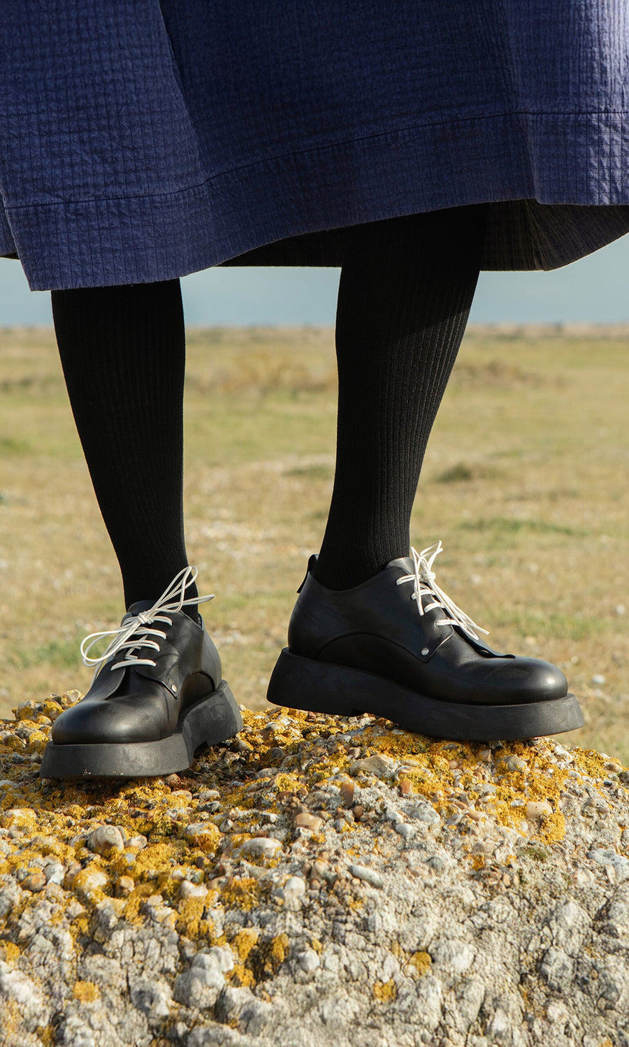 Plümo- Person wearing Hosier Brogues and black tights stands on a rock in a navy skirt in an outdoor grassy area.
