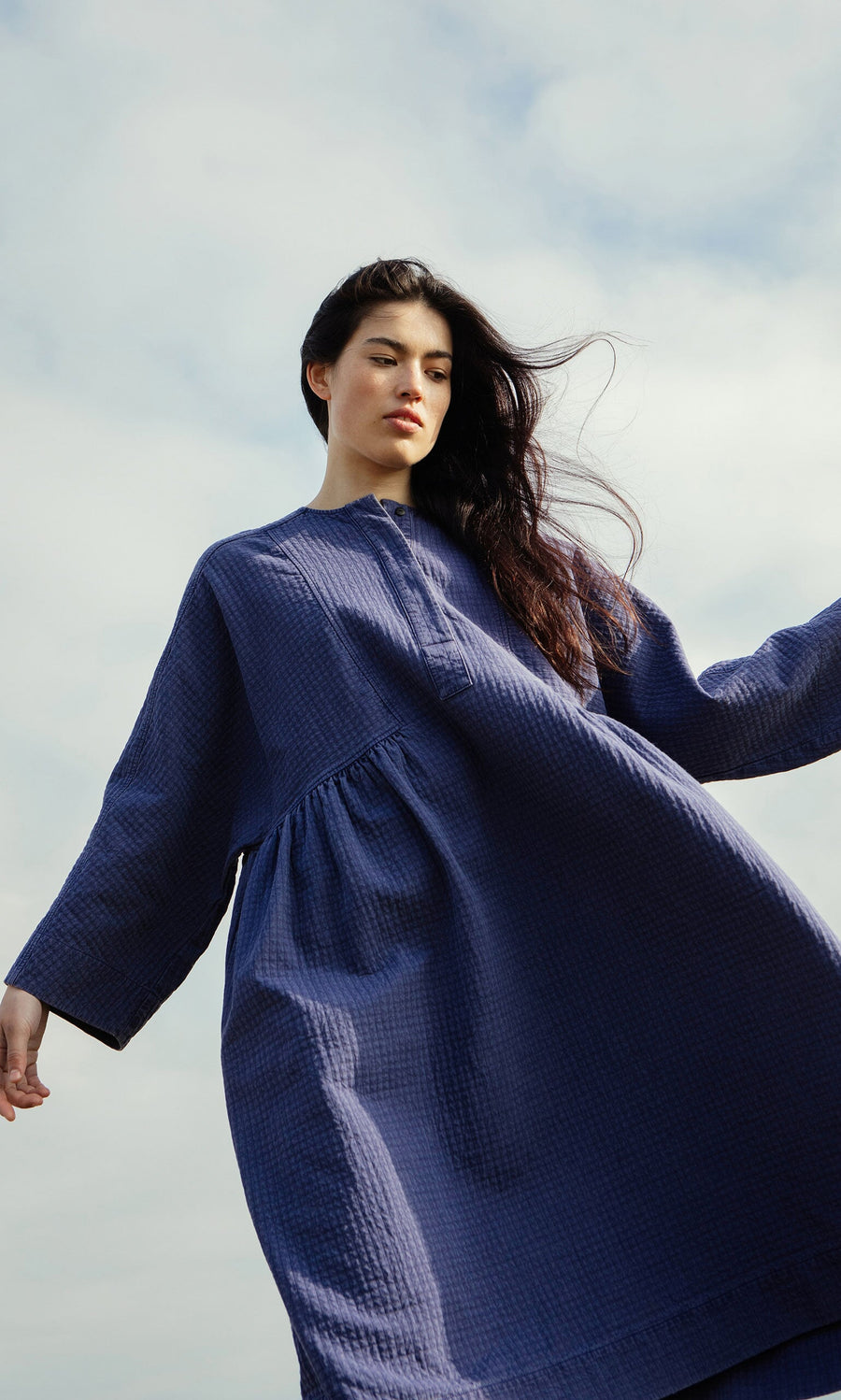Plümo- Woman wearing the Matilda Dress with a full skirt stands outdoors, her hair blowing in the wind under a cloudy sky.