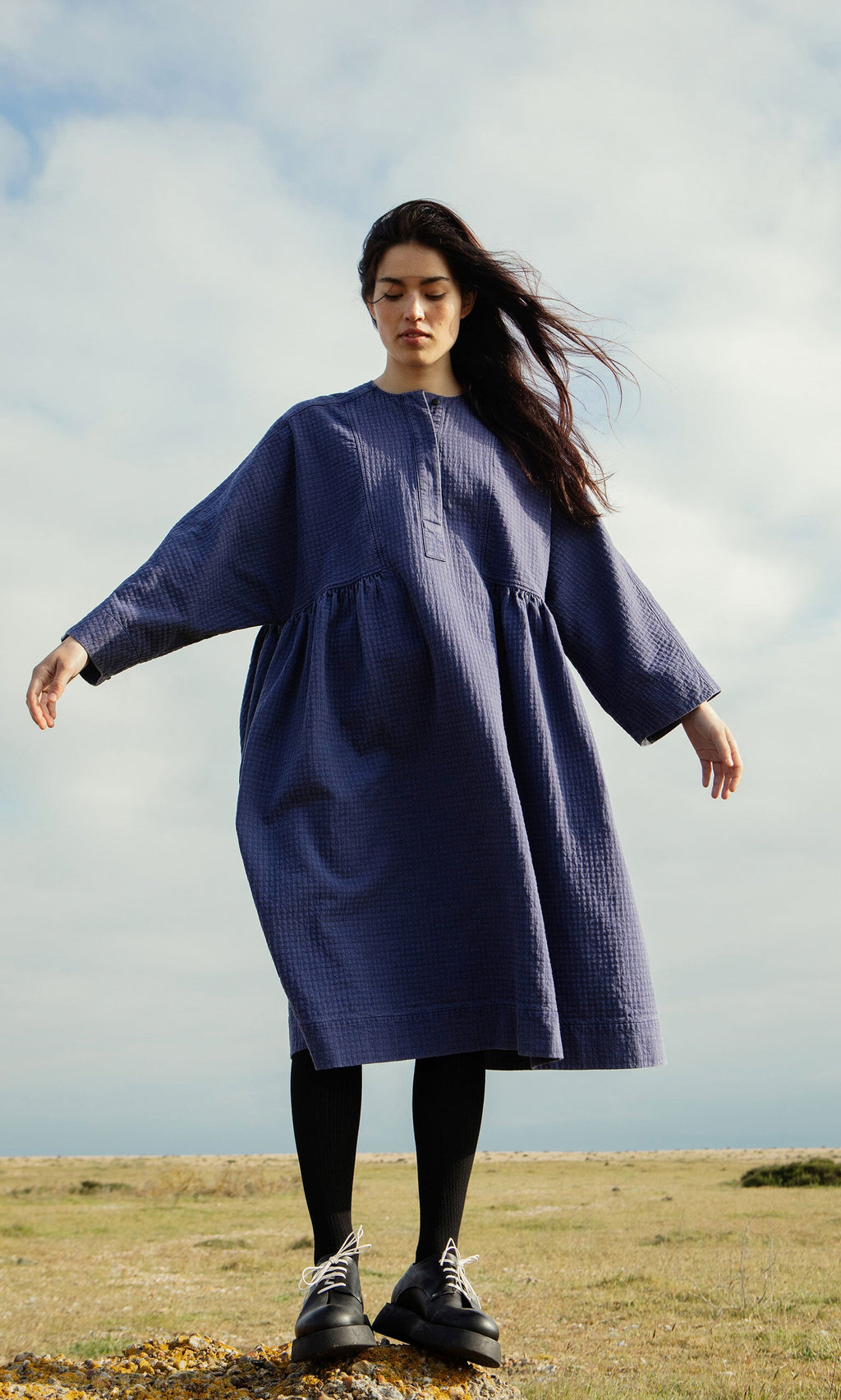 Plümo- Woman in the Matilda Dress, a loose long-sleeved indigo style with full skirt, stands outside on a cloudy day.