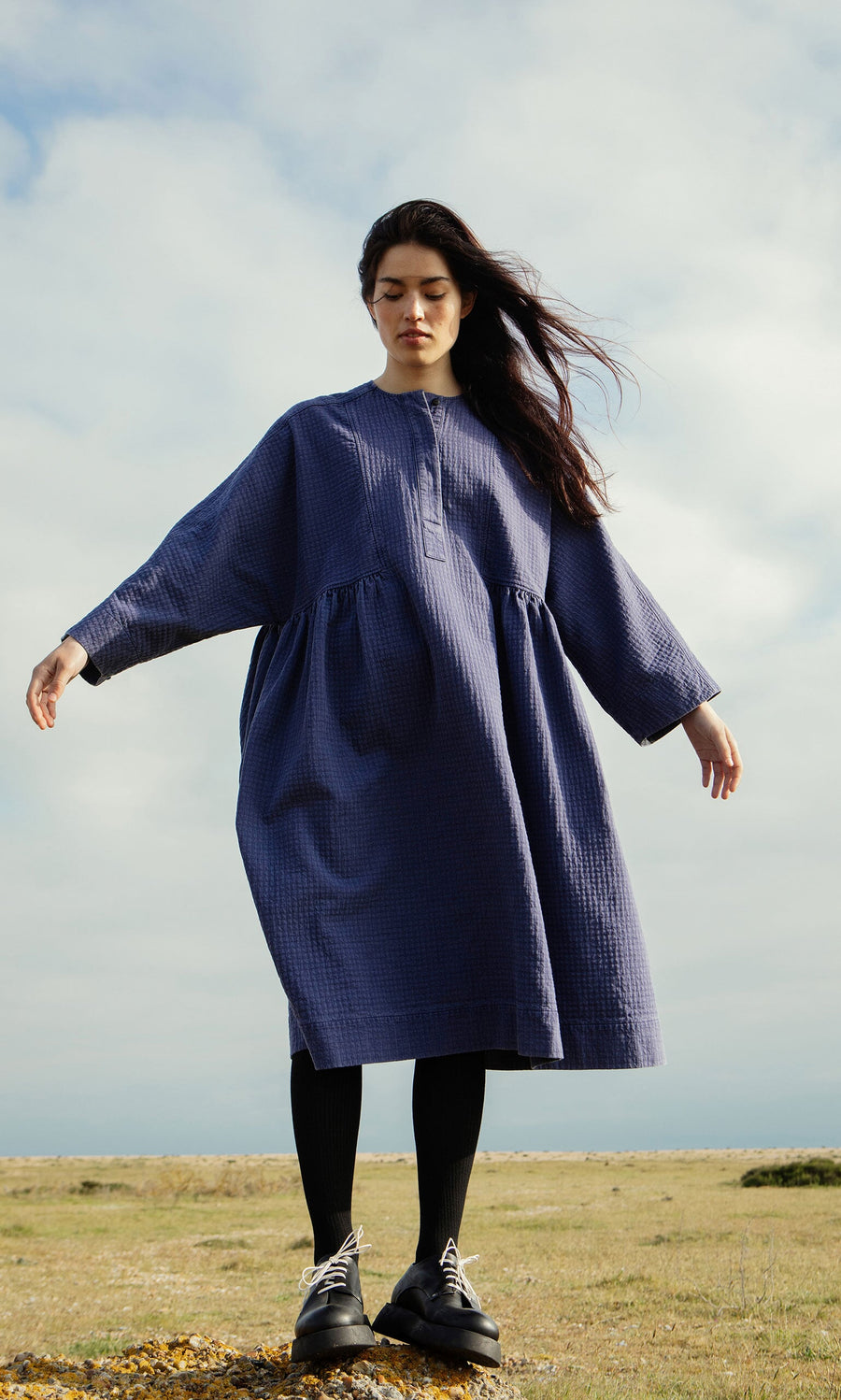 Plümo- Woman in the Matilda Dress, a loose long-sleeved indigo style with full skirt, stands outside on a cloudy day.