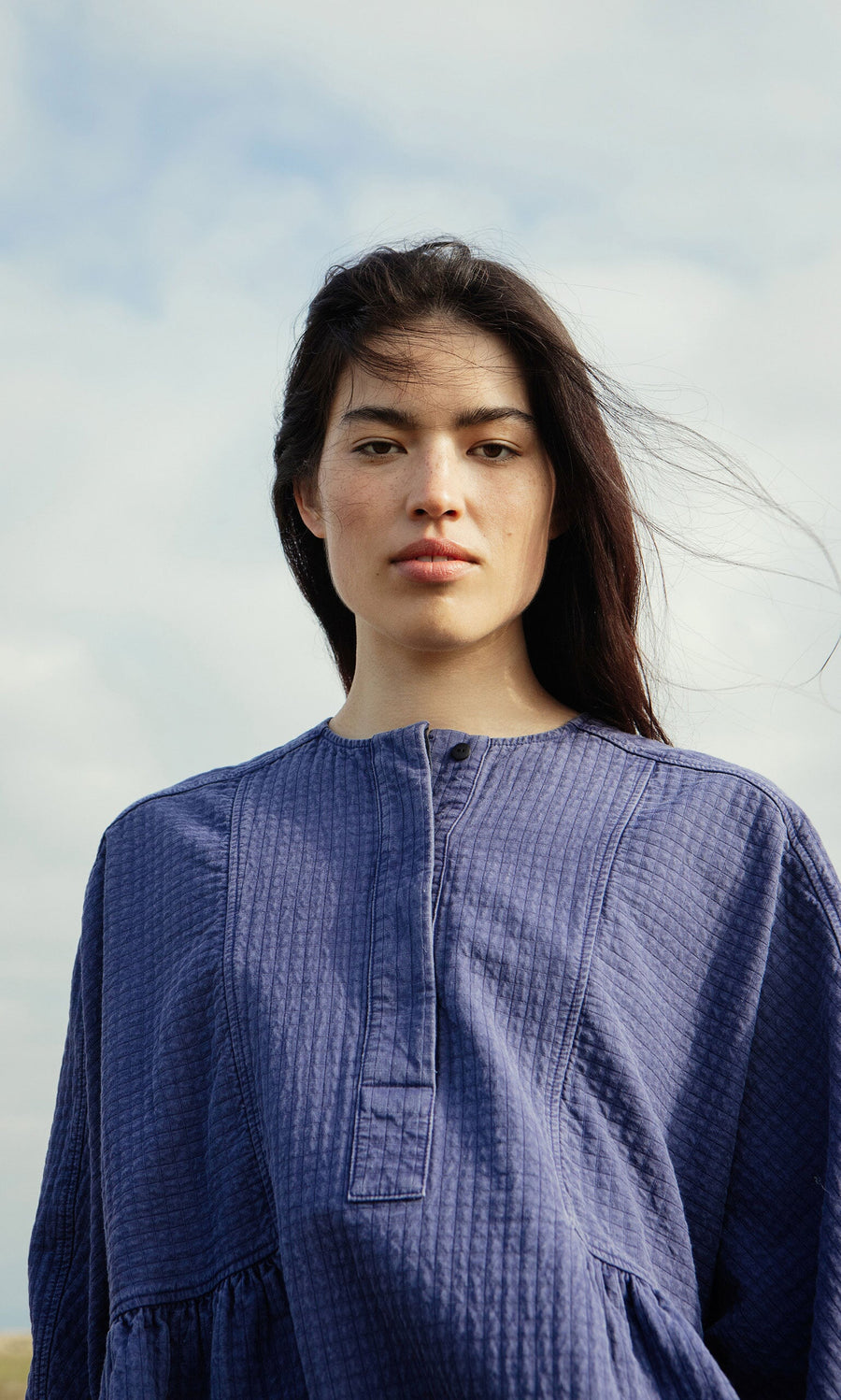 Plümo- Woman with long dark hair wears the Matilda Dress and a textured blue top outdoors under a cloudy sky, facing the camera.